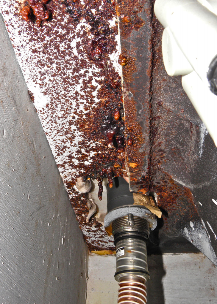Severe corrosion on metal surface near pipe connection, showing rust buildup and material degradation.