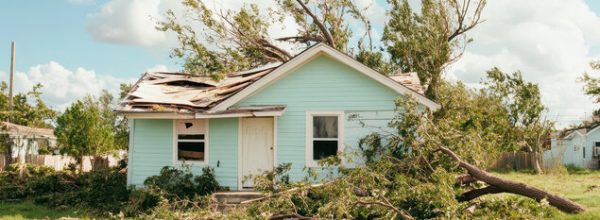 Small blue house with severe roof damage and fallen trees after a strong windstorm, illustrating the importance of wind-resistant construction.